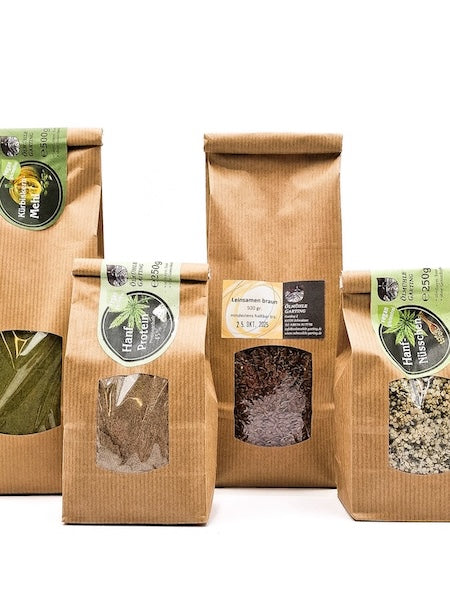 Vier braune Papiertüten mit Sichtfenster, gefüllt mit regionalen Produkten der Ölmühle Garting: Hanfmehl, Hanfprotein, Leinsamen, Hanfnüsse und Hanfschrot. Die Verpackungen stehen nebeneinander auf weißem Hintergrund.